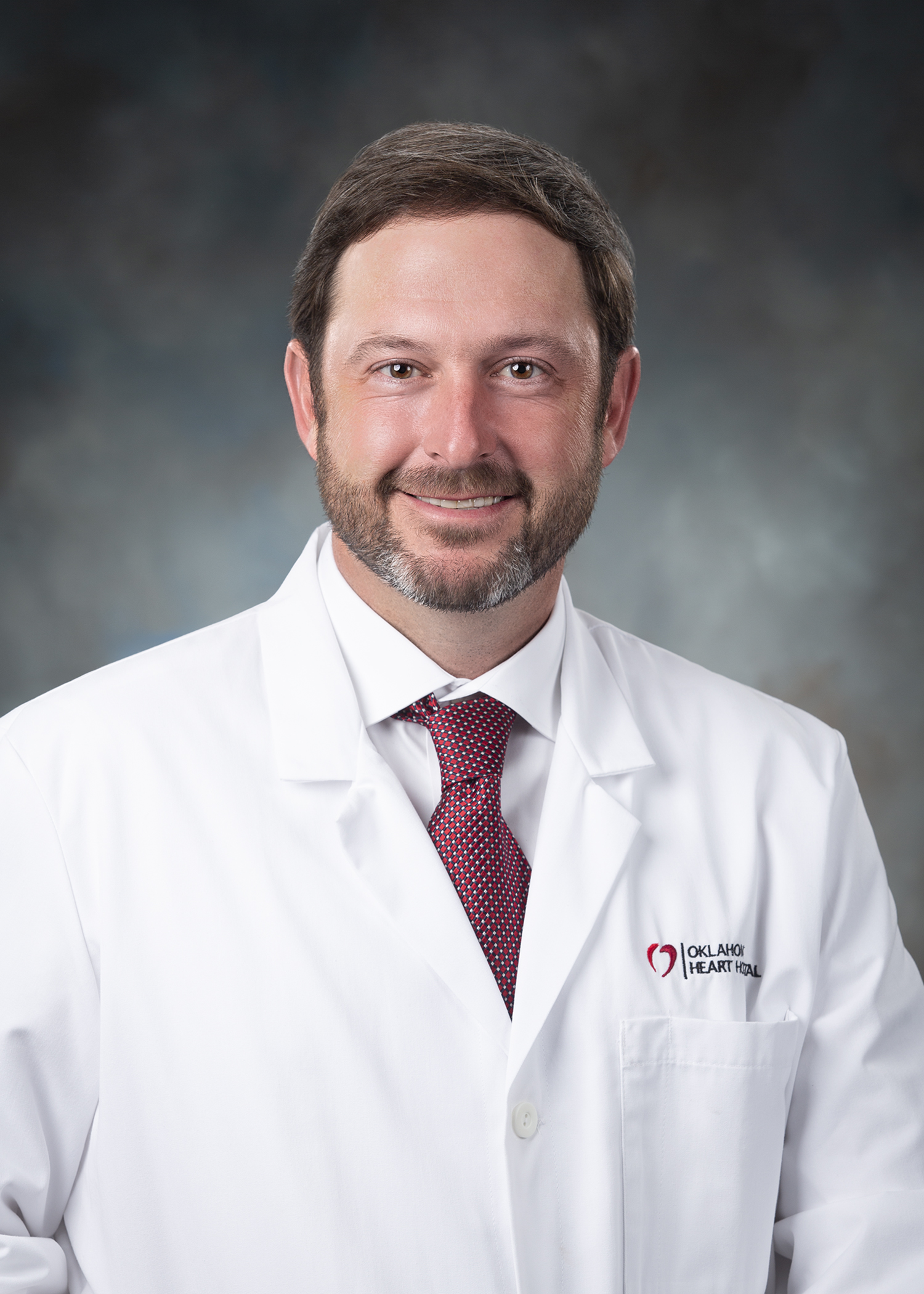 Dr. Beau Hawkins is wearing a white doctor's coat and red tie. He has short brown hair, brown beard and mustache.
