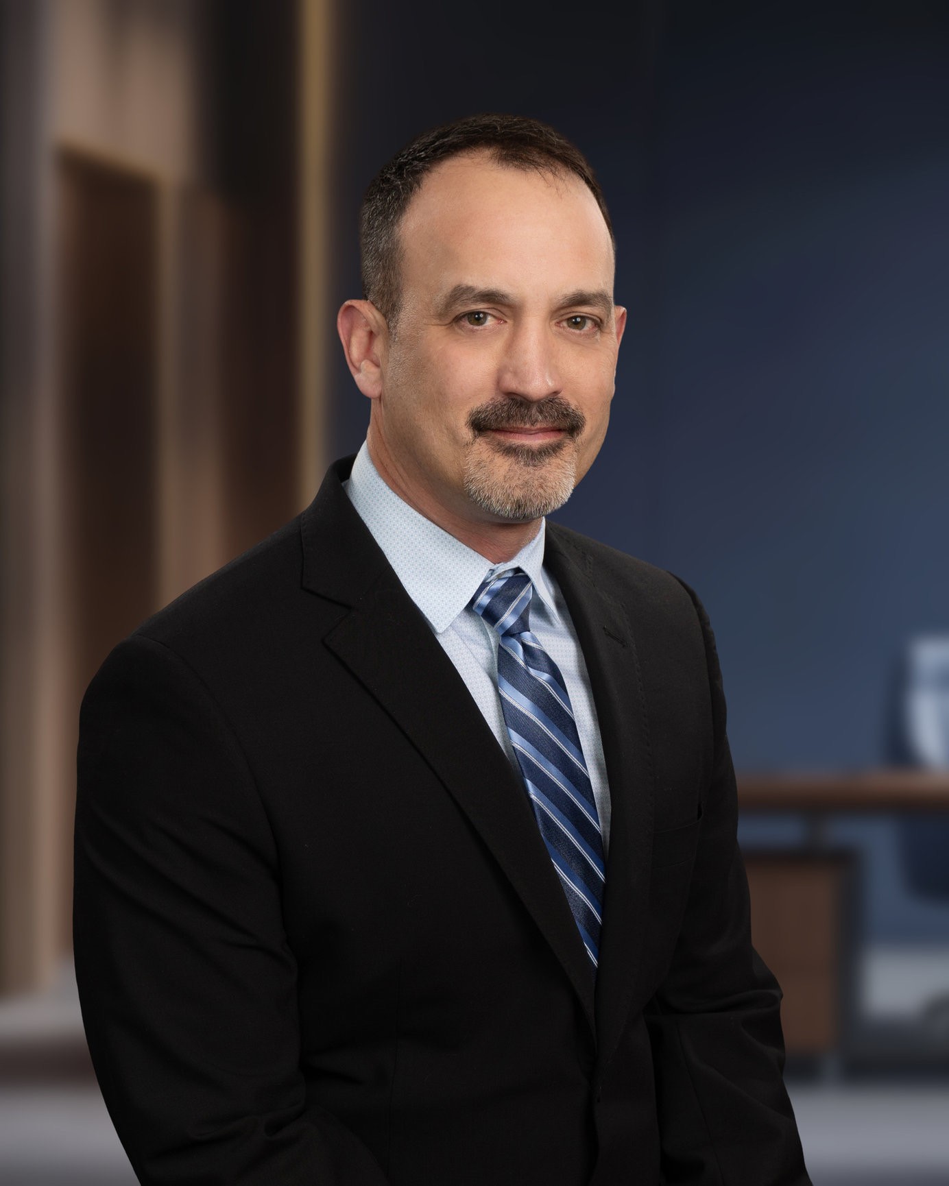 James Pritchett is wearing a black suit with blue tie. He has short brown hair and a mustache.