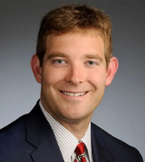 Cole Wootton, MD, pictured wearing a black suit and red tie. He has red-brown hair.